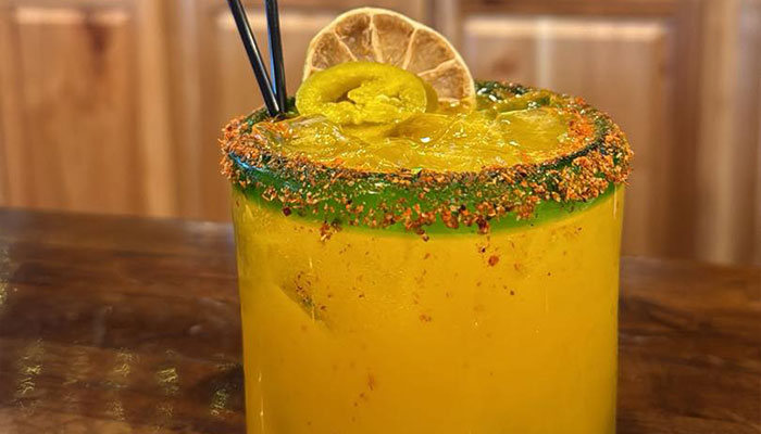 Yellow cocktail with ice, chili rim, jalapeño, lime slice garnish, and two black straws.