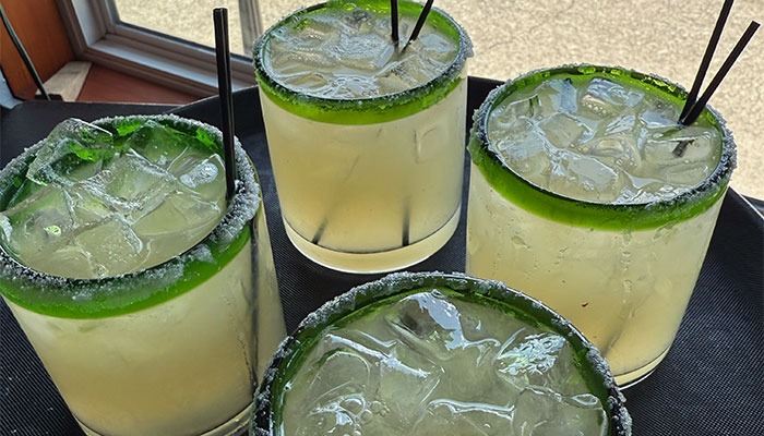 Four margaritas with salted rims and lime slices on a tray near a window.